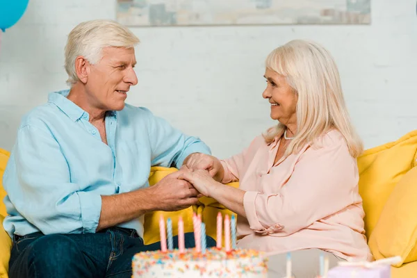 Feliz marido e mulher sênior sentado perto de bolo de aniversário, olhando um para o outro e de mãos dadas — Fotografia de Stock