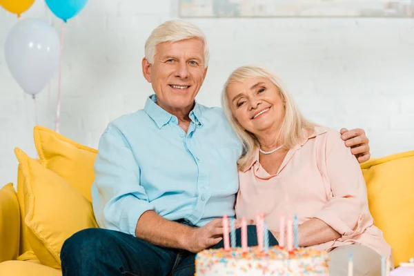 Feliz anciano marido y esposa sentado cerca de pastel de cumpleaños y sonriendo a la cámara - foto de stock
