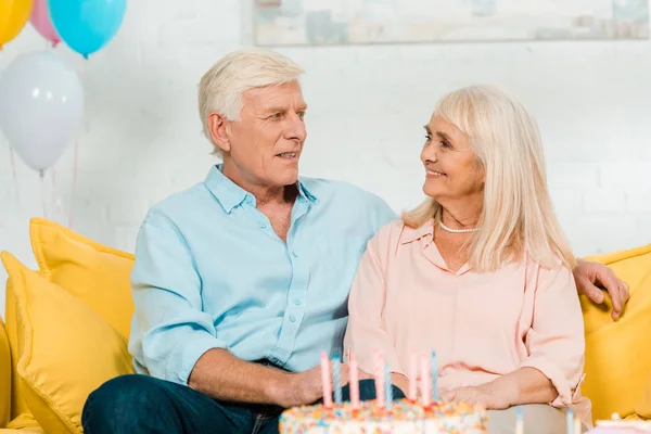 Happy senior husband and wife sitting near birthday cake and smiling at each other — Stock Photo