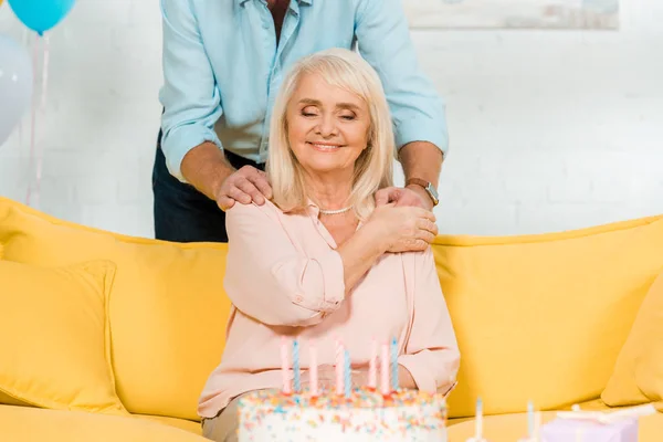 Vue recadrée de l'homme touchant les épaules de heureuse femme âgée assise près du gâteau d'anniversaire avec les yeux fermés — Photo de stock
