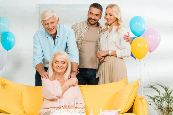 Heureuse femme âgée assise sur un canapé jaune près du gâteau d'anniversaire et la famille souriante — Photo de stock