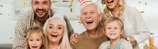 Panoramic shot of happy family looking at camera under falling confetti — Stock Photo