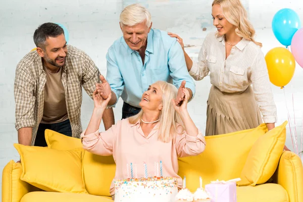 Mulher sênior feliz sentado no sofá amarelo perto de bolo de aniversário e família sorridente — Fotografia de Stock
