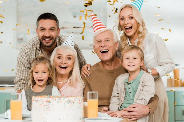 Glückliche Familie blickt in die Kamera, während sie neben Geburtstagstorte unter fallendem Konfetti sitzt — Stockfoto