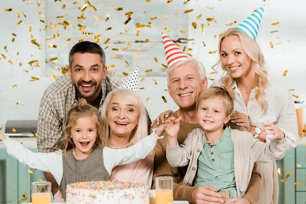 Happy family sitting under falling confetti near birthday cake and smiling at camera — Stock Photo