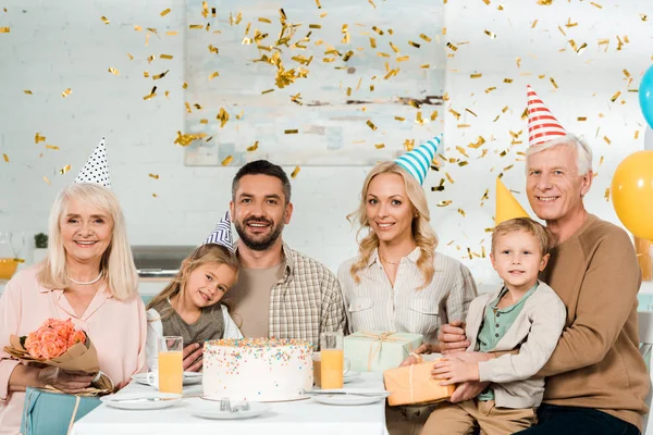 Fröhliche Familie sitzt am Küchentisch neben Geburtstagstorte unter fallendem Konfetti und lächelt in die Kamera — Stockfoto