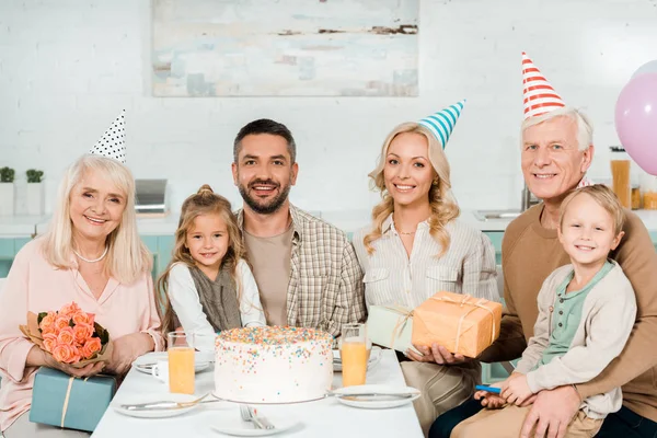 Familia alegre sentado en la mesa de la cocina cerca de pastel de cumpleaños y mirando a la cámara - foto de stock