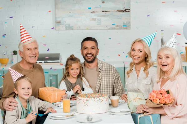 Glückliche Familie sitzt am Küchentisch neben Geburtstagstorte unter fallendem Konfetti — Stockfoto