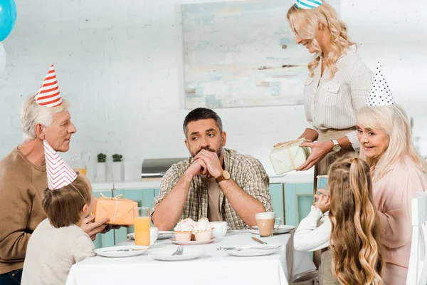 Hombre mayor y mujer adulta sosteniendo cajas de regalo cerca de hombre pensativo sentado en la mesa de la cocina con la familia - foto de stock