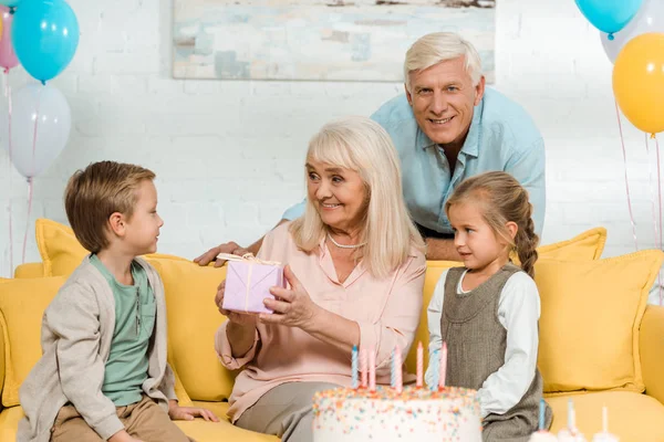 Happy senior woman holding gift box while sitting on yellow sofa near grandchildren and husband — Stock Photo