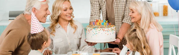 Обрізаний вид чоловіка, що тримає торт на день народження зі свічками біля щасливої сім'ї, що сидить за кухонним столом — стокове фото