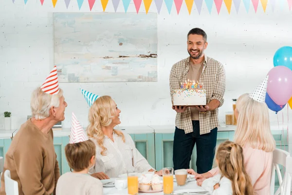 Hombre sonriente sosteniendo pastel de cumpleaños con velas cerca de la familia feliz sentado en la mesa de la cocina - foto de stock