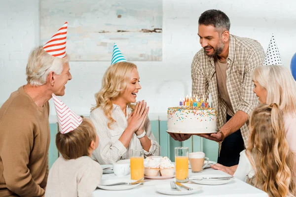 Hombre sonriente sosteniendo pastel de cumpleaños con velas cerca de la familia feliz sentado en la mesa de la cocina - foto de stock