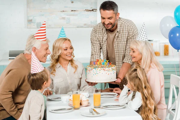 Hombre feliz celebración de pasteles de cumpleaños con velas cerca de la familia sentado en la mesa de la cocina - foto de stock