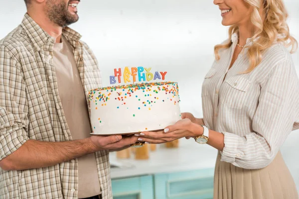 Cropped view of smiling husband and wife holding birthday cake with colorful candles and happy birthday lettering — Stock Photo