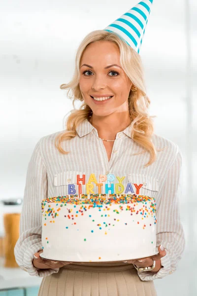 Happy woman in party cap holding birthday cake with colorful candles and happy birthday lettering — Stock Photo