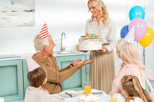 Mujer sonriente presentando pastel de cumpleaños al hombre mayor sentado en la mesa de la cocina cerca de la familia - foto de stock