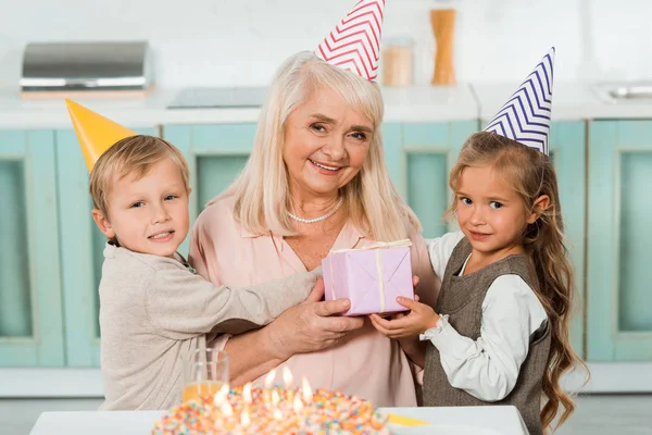 Cute kids hugging happy grandmother holding gift box while sitting near birthday cake — Stock Photo