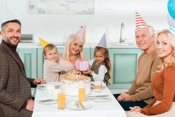 Niños alegres sentados en vueltas de la abuela mientras se sienta cerca de pastel de cumpleaños con la familia - foto de stock