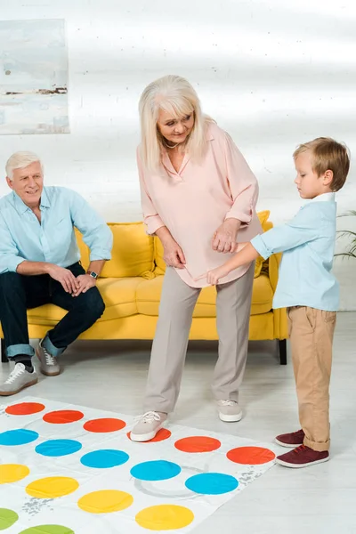 Kyiv, Ukraine - 21. November 2019: Lächelnde Oma und Enkel spielen Twister-Spiel in der Nähe des Großvaters, der auf dem Sofa sitzt — Stockfoto