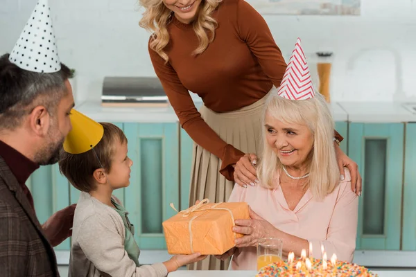 Обрезанный вид женщины, трогающей плечи счастливой бабушки, забирающей подарочную коробку у внука — стоковое фото