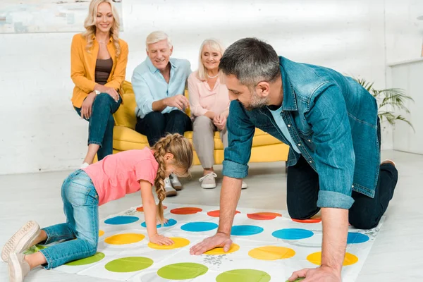 KYIV, UCRAINA - 21 NOVEMBRE 2019: padre e figlia allegri che giocano a twister game vicino alla famiglia seduti sul divano — Foto stock