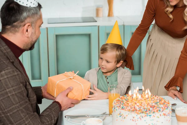 Padre presentando caja de regalo a hijo feliz mientras está sentado en la mesa de la cocina cerca de pastel de cumpleaños - foto de stock