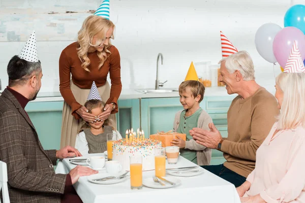 Усміхнена жінка покриває дочки очима руками, а вся сім'я сидить біля торта на день народження — стокове фото