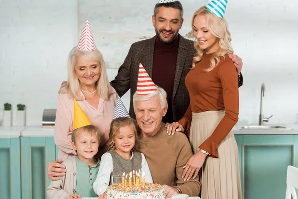 Happy family in party caps looking at birthday cake with burning candles — Stock Photo