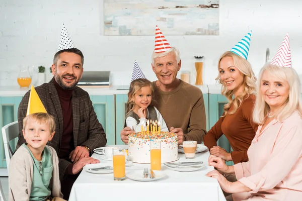 Happy family in party caps smiling at camera while sitting near birthday cake — Stock Photo