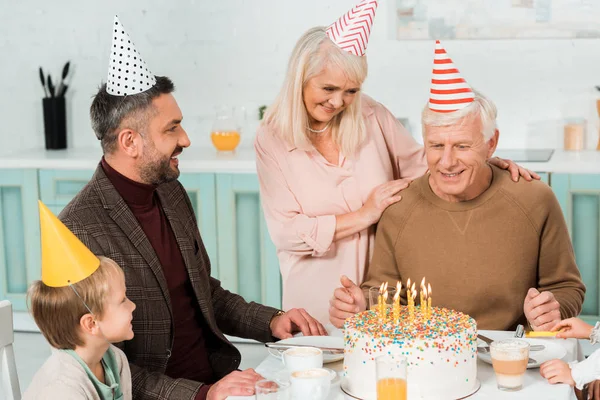 Senior woman touching shoulders of happy man sitting near birthday cake with family — Stock Photo