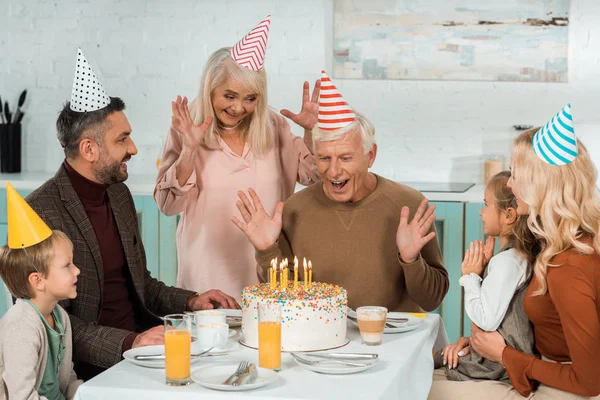 Excited senior man looking at birthday cake with burning candles while sitting at served table near family — Stock Photo