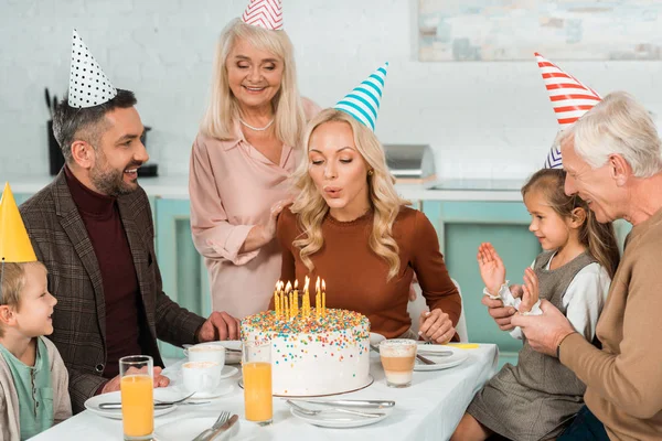 Femme heureuse soufflant des bougies brûlantes sur le gâteau d'anniversaire près de la famille assis à la table de cuisine — Photo de stock