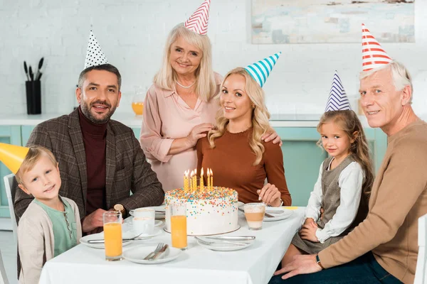 Glückliche Familie lächelt in die Kamera, während sie am Küchentisch neben Geburtstagstorte sitzt — Stockfoto