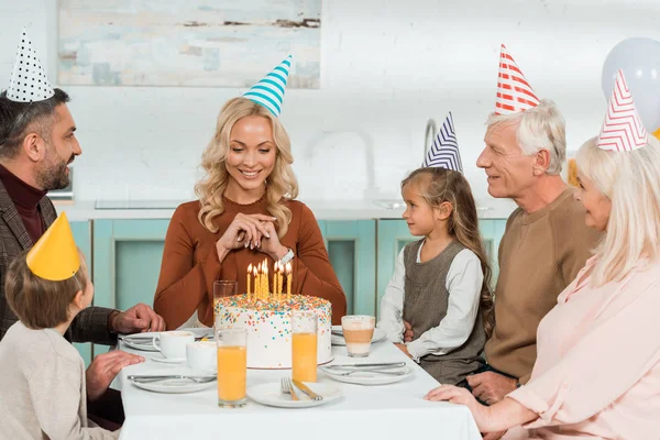 Щаслива жінка дивиться на торт на день народження, сидячи за кухонним столом з сім'єю — стокове фото
