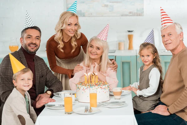 Улыбающаяся женщина касается плеч счастливой пожилой женщины, в то время как вся семья сидит возле торта день рождения — стоковое фото