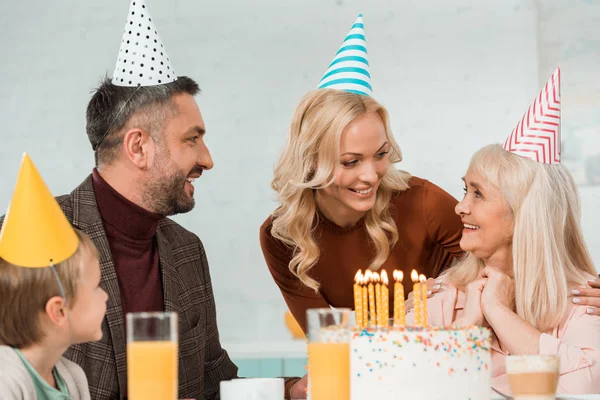 Весела сім'я, дивлячись на щасливу старшу жінку, що сидить біля торта на день народження — стокове фото
