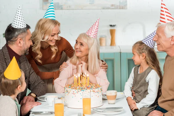 Donna sorridente toccare le spalle della donna anziana felice mentre tutta la famiglia seduta vicino torta di compleanno — Foto stock