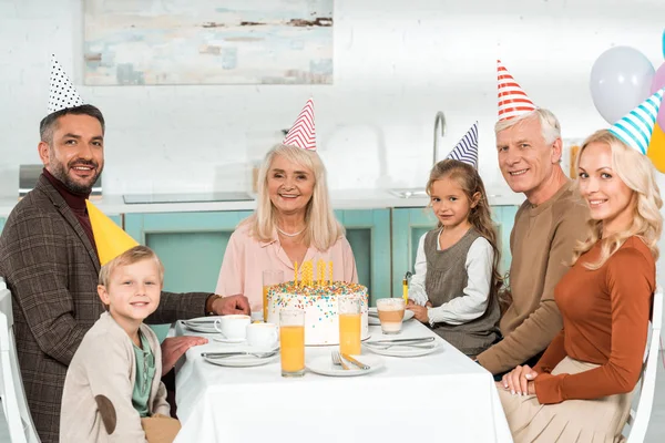 Família feliz olhando para a câmera enquanto sentado perto da mesa da cozinha com bolo de aniversário — Fotografia de Stock