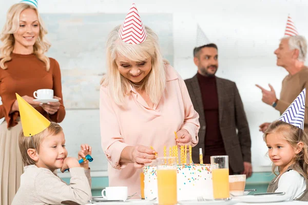 Счастливая пожилая женщина кладет свечи в торт на день рождения рядом с семьей в партии колпачки — стоковое фото