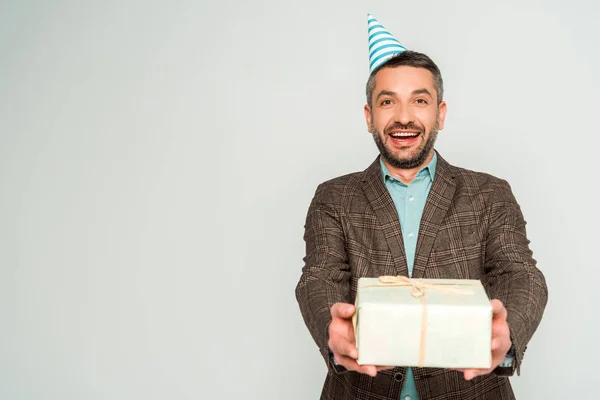 Felice uomo in cappello partito in possesso di scatola regalo, mentre guardando la fotocamera isolata su grigio — Foto stock