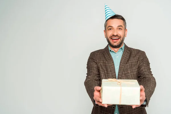 Hombre feliz en la tapa del partido sosteniendo caja de regalo mientras mira la cámara aislada en gris - foto de stock