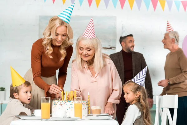 Mujeres felices poniendo velas en pastel de cumpleaños cerca de la familia en gorras de fiesta - foto de stock