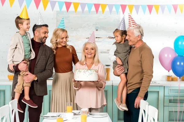 Happy senior woman holding birthday cake near cheerful family — Stock Photo