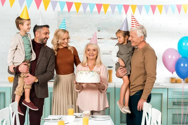 Mulher sênior feliz segurando bolo de aniversário perto da família alegre — Fotografia de Stock