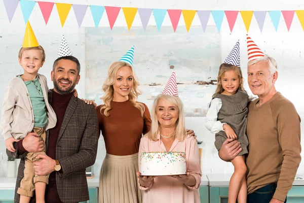 Happy senior woman holding birthday cake near family in party hats smiling at camera — Stock Photo
