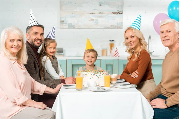 Família feliz sentado na mesa de cozinha servida com bolo de aniversário e olhando para a câmera — Fotografia de Stock