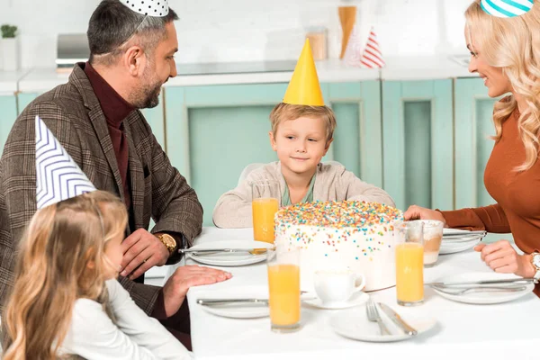 Cute kid looking at birthday cake while sitting at served table near parents and sister — Stock Photo