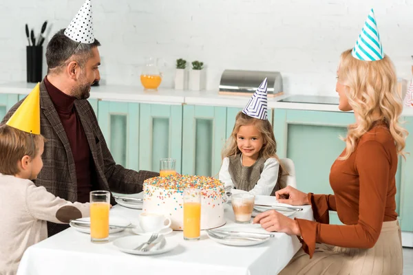 Lindo niño mirando pastel de cumpleaños mientras está sentado en la mesa servida cerca de los padres y el hermano - foto de stock