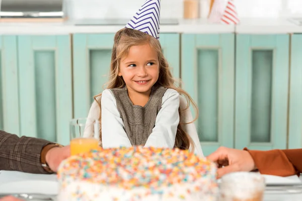 Selektiver Fokus des glücklichen Kindes in Partymütze, das am Küchentisch neben der Geburtstagstorte sitzt — Stockfoto